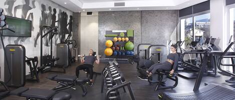 A modern gym with various exercise equipment, a wall-mounted TV, and a large window offering a city view.