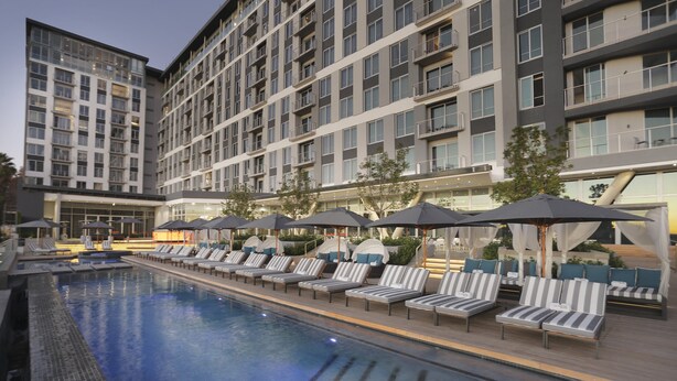 Une piscine entourée de chaises longues et de parasols, elle-même bordée d’un immeuble à plusieurs étages doté de balcons.