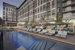 Una piscina con sillones de descanso y sombrillas, rodeada por un edificio de varios pisos con balcones.
