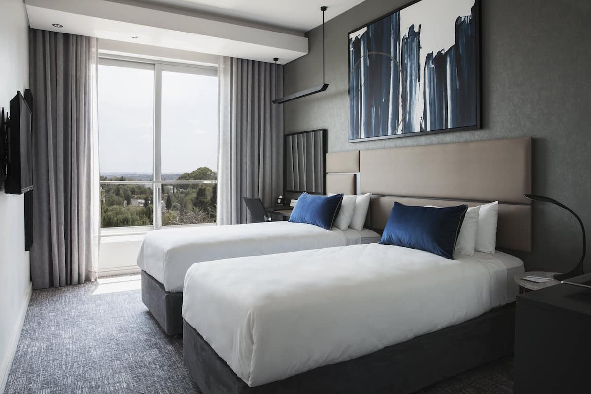 Habitación de hotel con dos camas, un ventanal con cortinas, un escritorio y una obra de arte colgada.