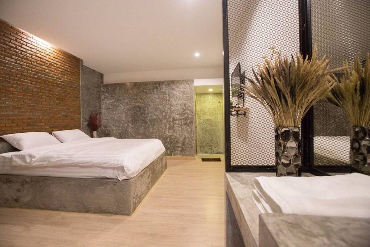 A modern bedroom with a brick accent wall, a large bed, a marble-textured wall, and a decorative vase with dried plants.