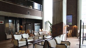 Um lobby amplo com pé-direito alto, parede de tijolos e uma grande janela. Há várias áreas de estar com cadeiras e mesas.