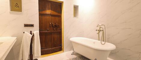 Une salle de bain avec une porte en bois, une baignoire sur pieds, un carrelage imitant le marbre et un lavabo blanc.