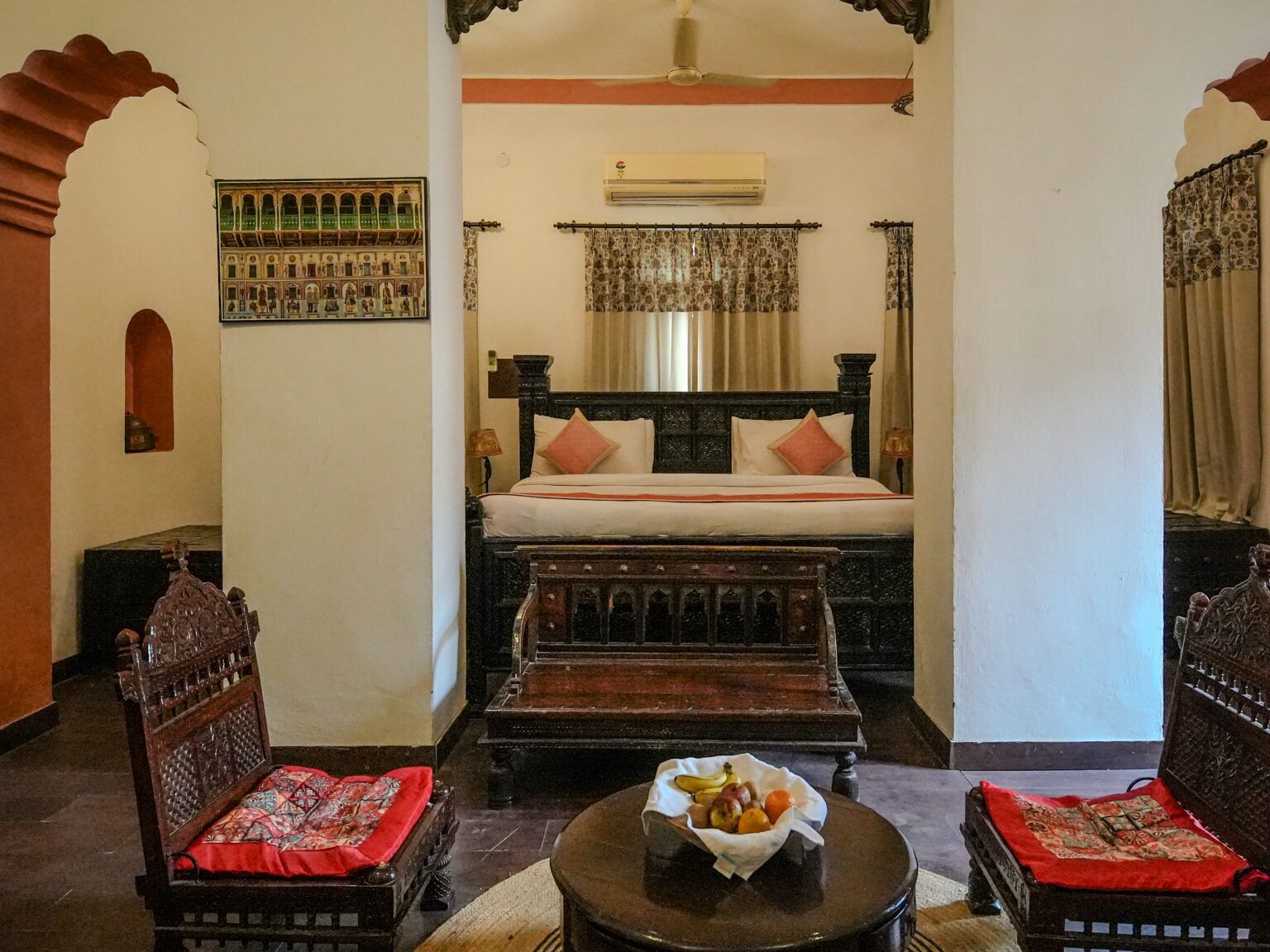 A traditional room with a bed, two wooden chairs, a small table with fruit, and a wall-mounted air conditioner.