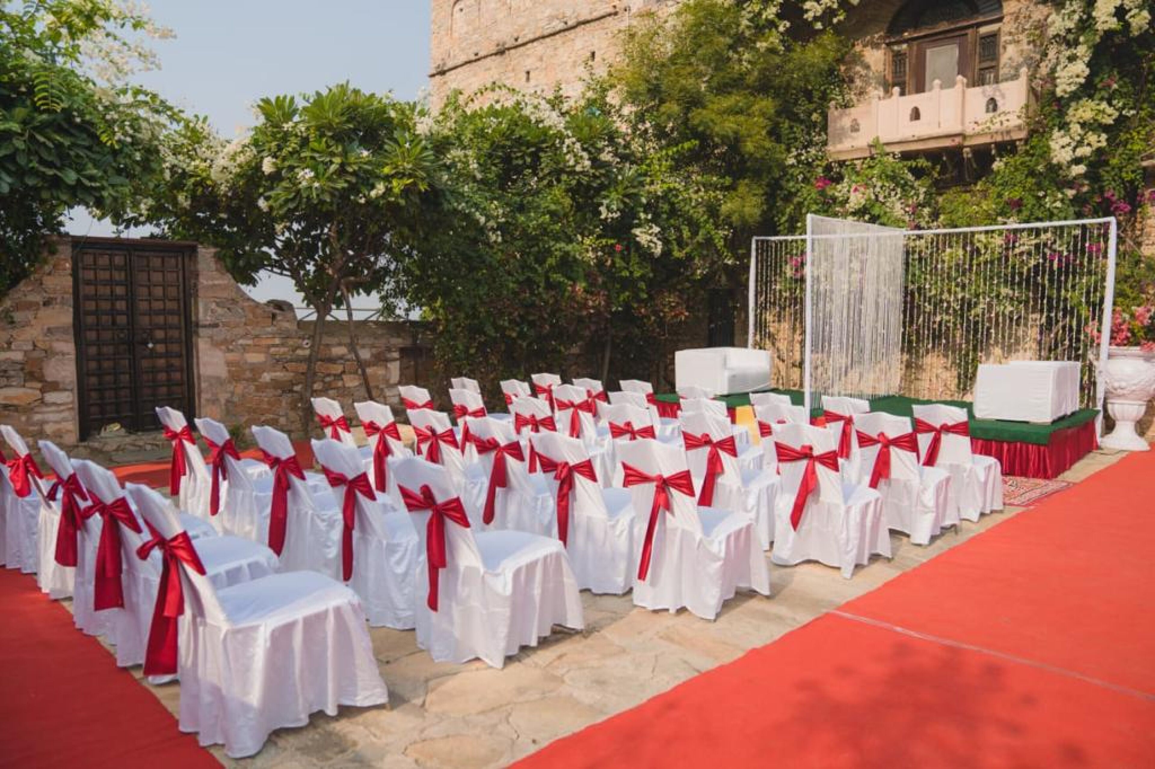 Evento con alfombra roja, filas de sillas blancas atadas con cintas rojas, frente a un muro de piedra y abundante vegetación.