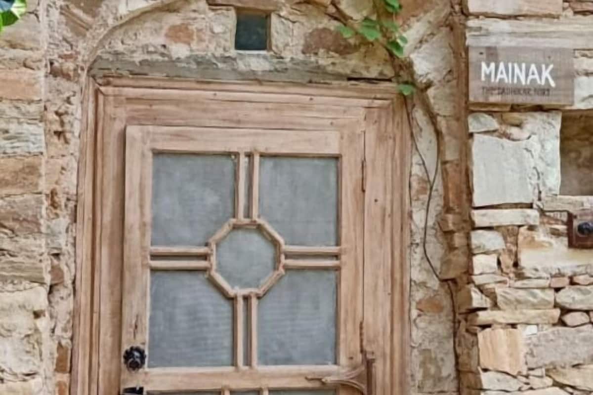 Ein rustikales Steinhaus mit einer Holztür, die ein geometrisches Glasspiegelmuster aufweist. Über der Tür befindet sich ein kleines Fenster und ein Schild mit der Aufschrift 'MAINAK'.