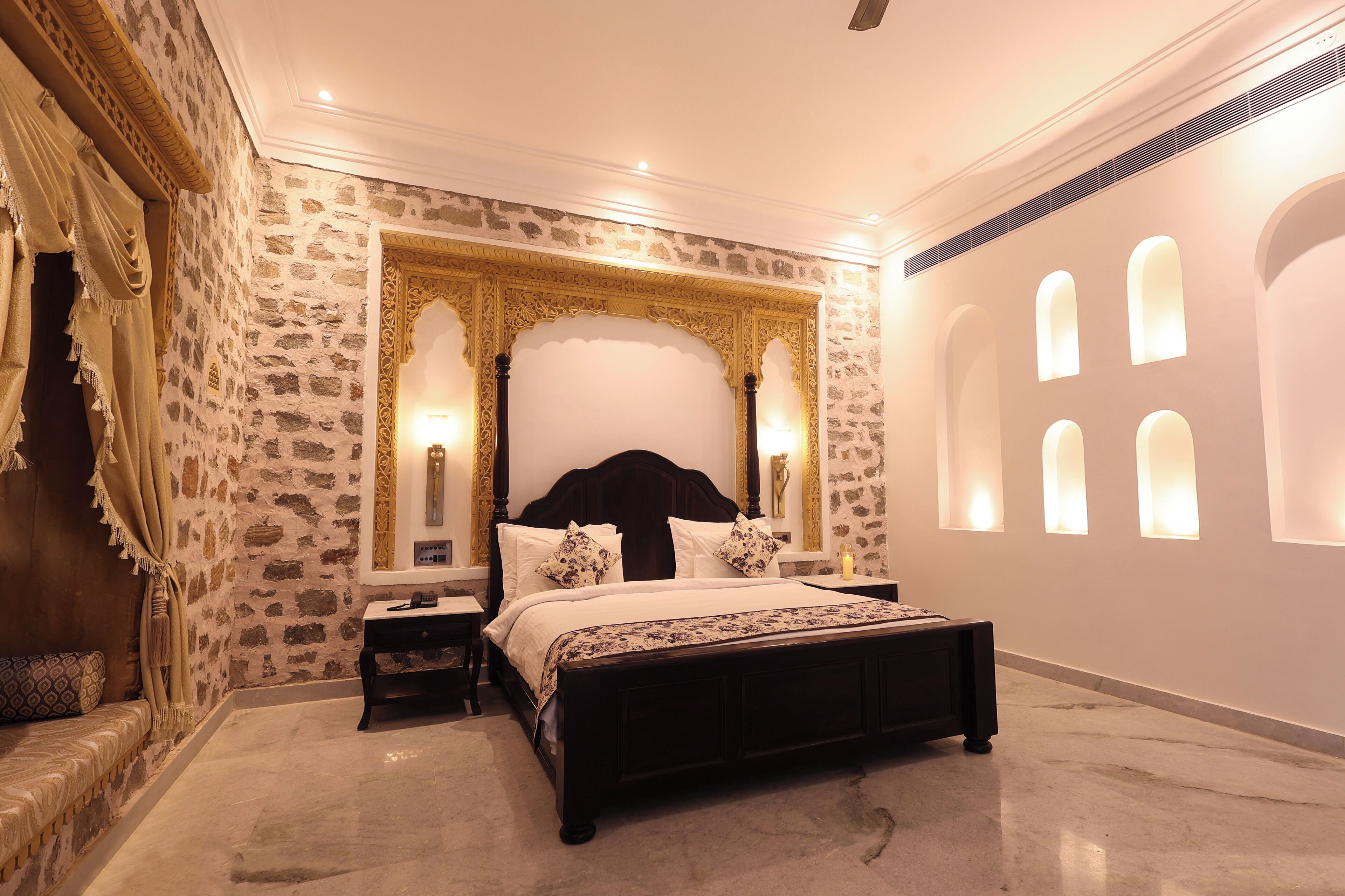 A bedroom with a stone wall, a large bed with a dark headboard, and a nightstand with a lamp.