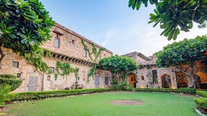 A courtyard with a central lawn, stone buildings with arched doorways, and lush greenery. - Fort Dadhikar, Alwar (Alwar)