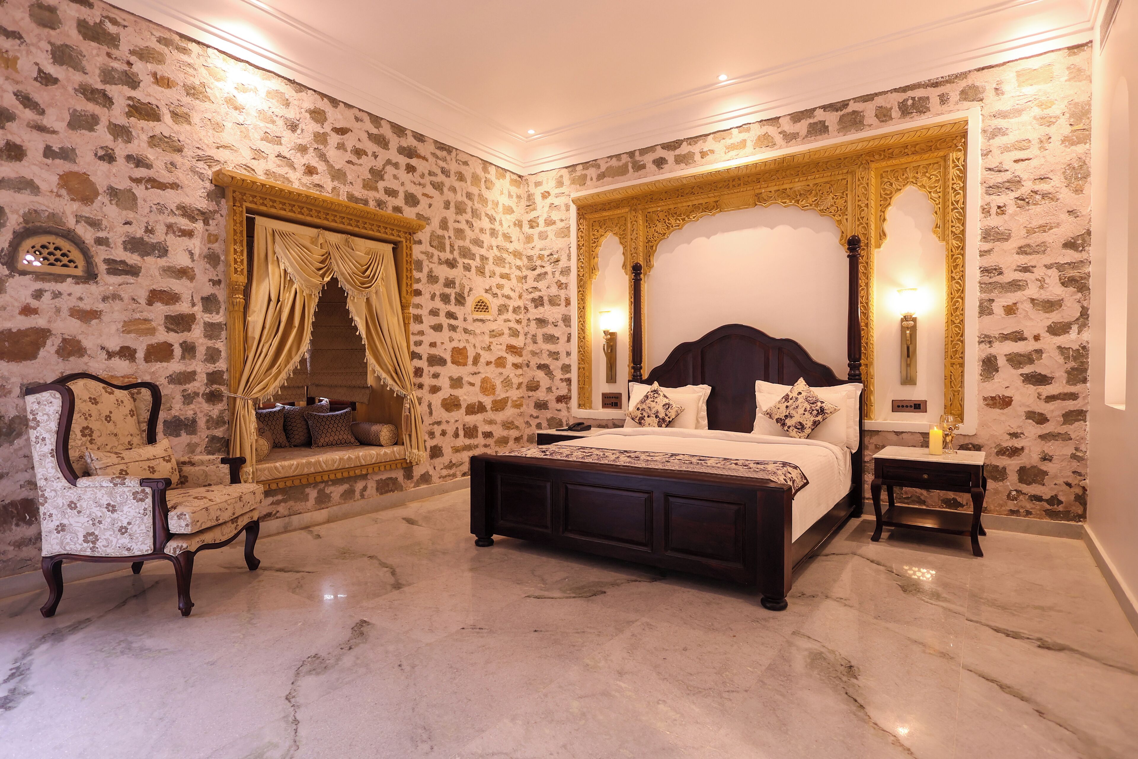 A bedroom with a stone wall, a large bed with a canopy, a chair, and a nightstand with a lamp.