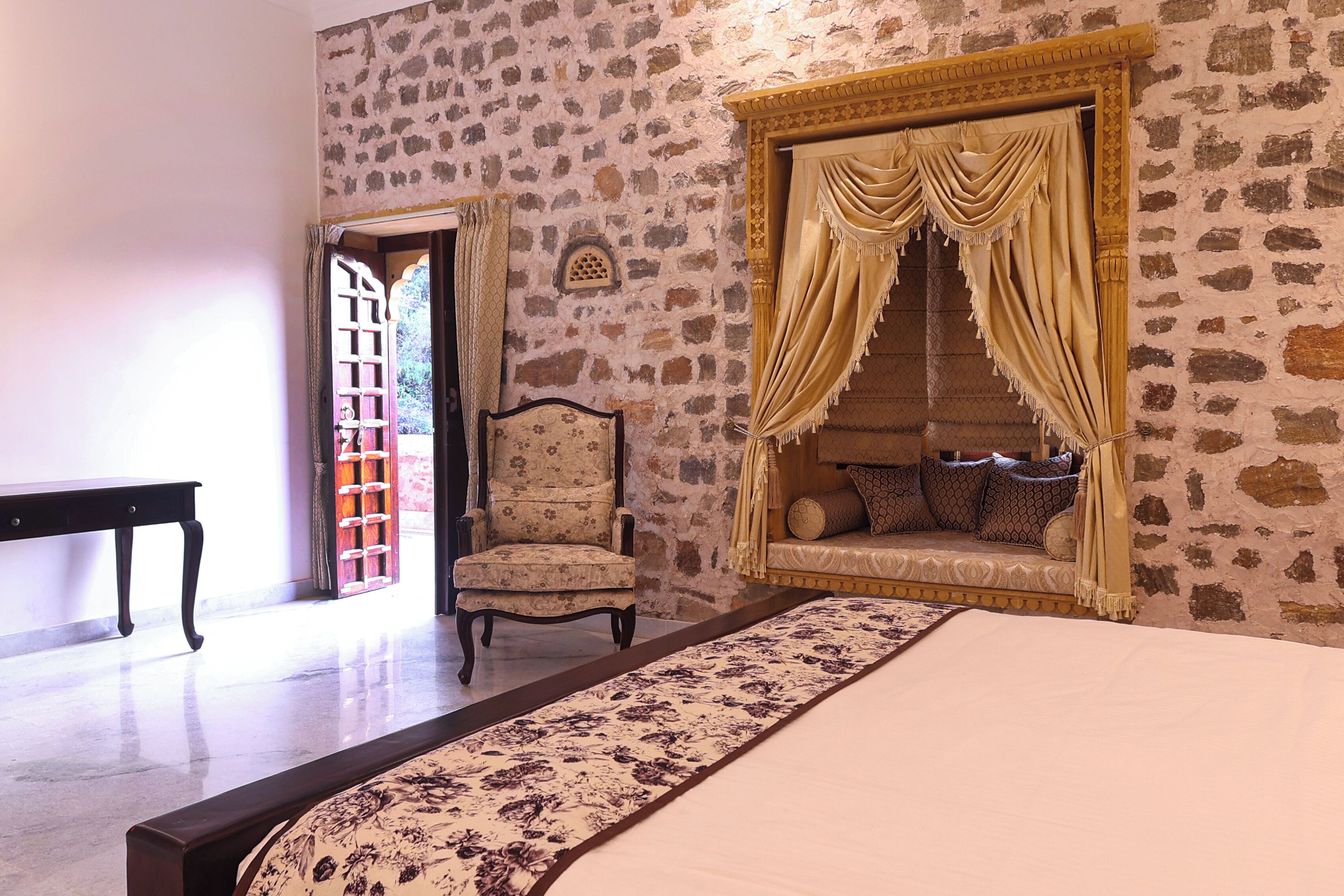 A bedroom with a stone wall, a bed with a floral bedspread, a chair, and a window with curtains.