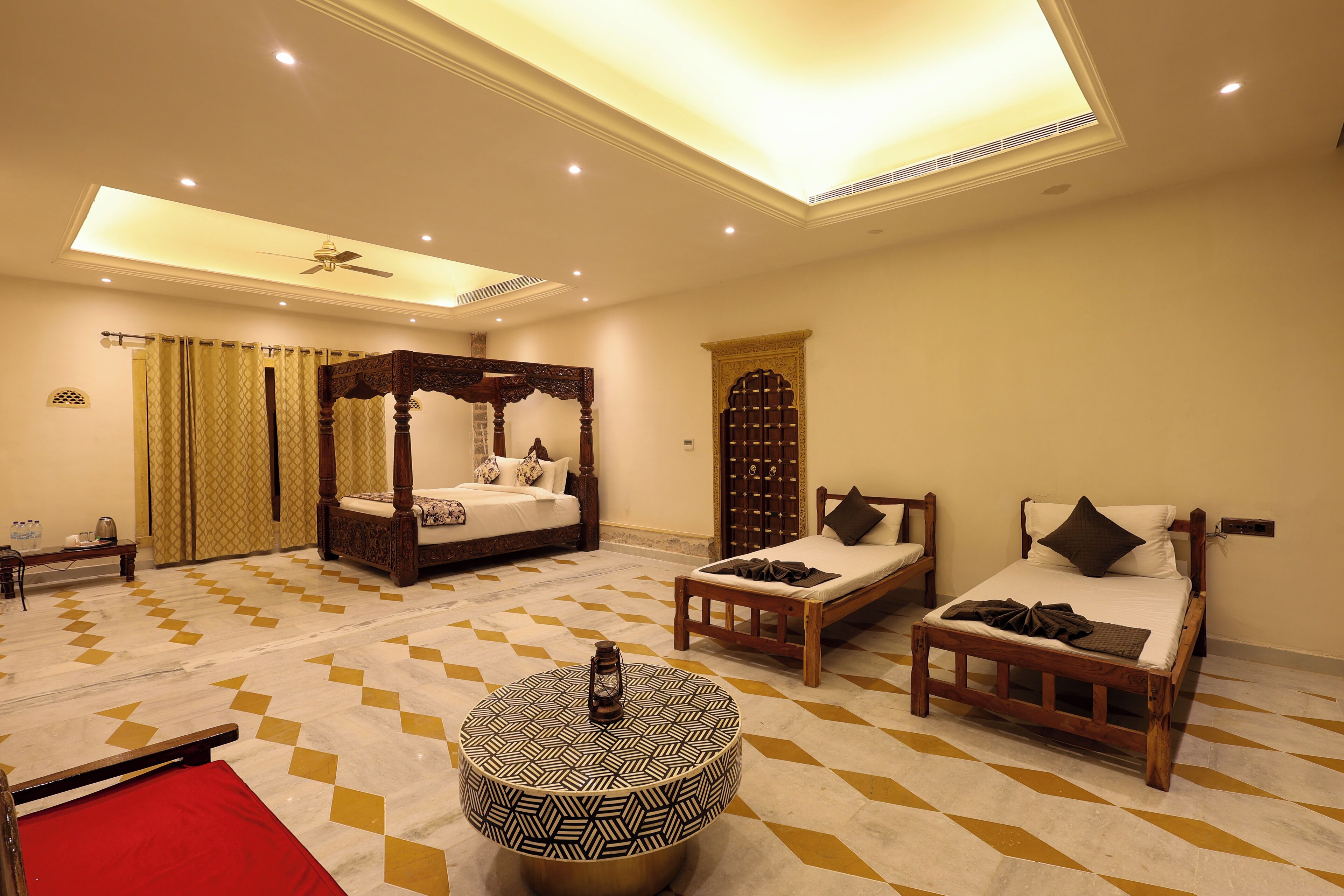 A hotel room with a large bed, two smaller beds, a round ottoman, a ceiling fan, and a patterned floor.