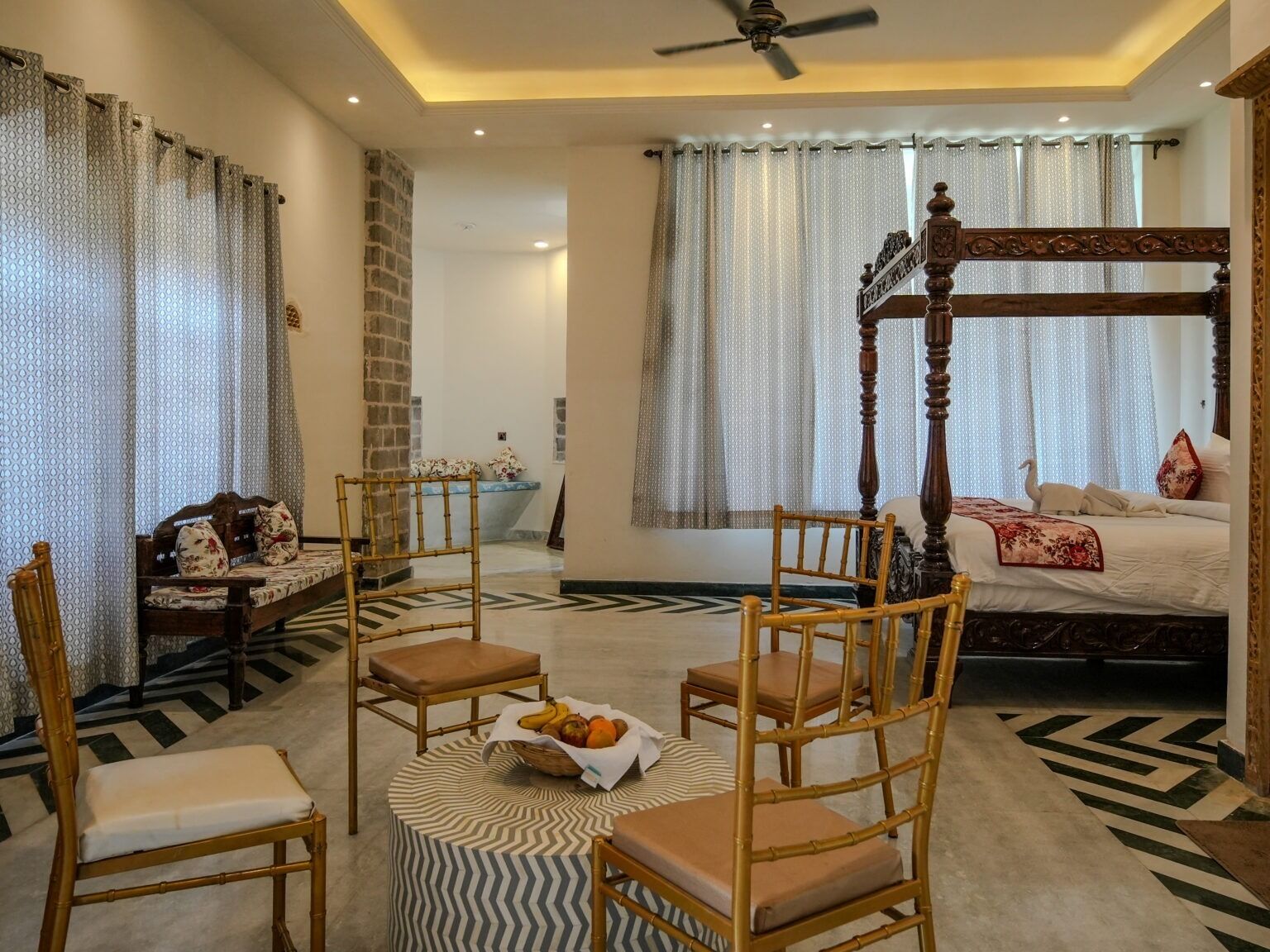 A bedroom with a four-poster bed, a sitting area with chairs, a small table with fruit, and a patterned rug.