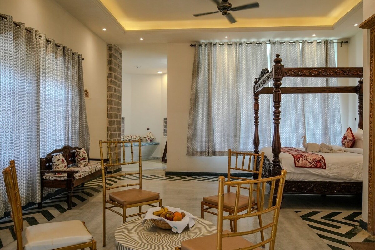A bedroom with a four-poster bed, a sitting area with chairs, a small table with fruit, and a patterned rug.
