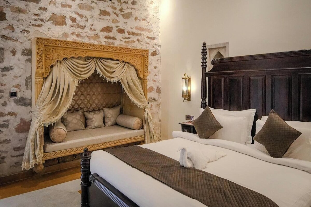 A bedroom with a stone wall, a canopy bed, a wooden headboard, and a bedside table with a lamp.