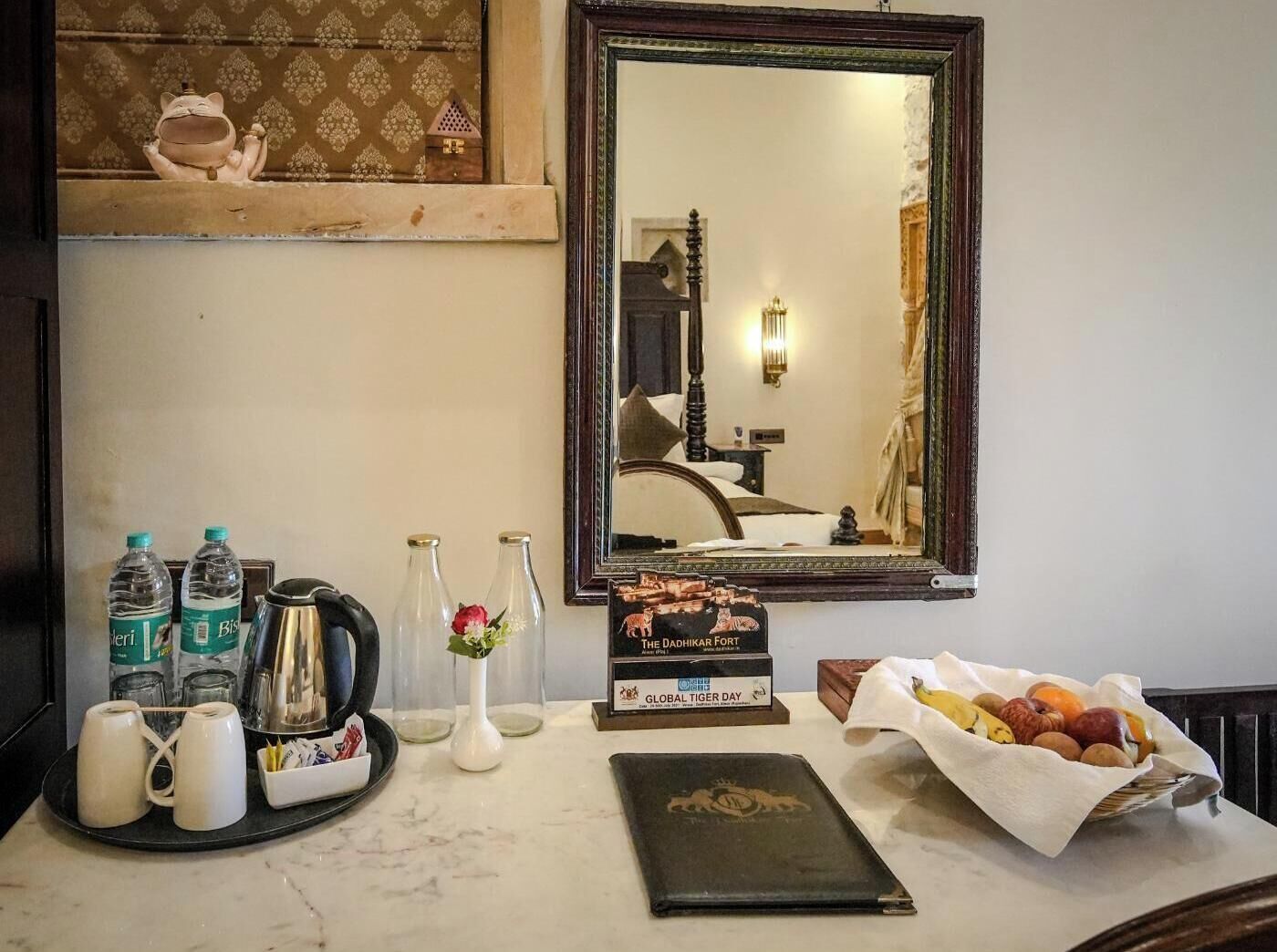 Une chambre d’hôtel comprenant un lit, un miroir, un plateau avec des bouteilles d’eau, une cafetière, un vase avec une fleur, un livre intitulé « Le fort de Dadhikar » et un panier de fruits.