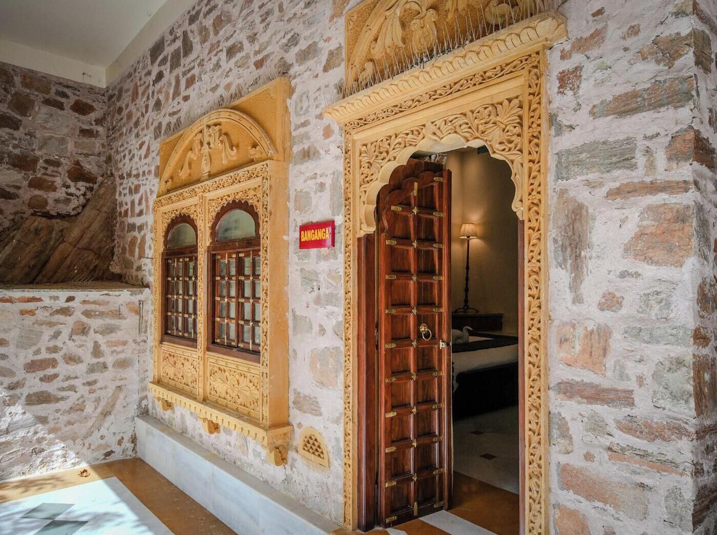 Une pièce aux murs de pierre, dotée de portes et de fenêtres en bois sculpté, une enseigne indiquant « BANGANGA », et un lit visible à travers une porte ouverte.