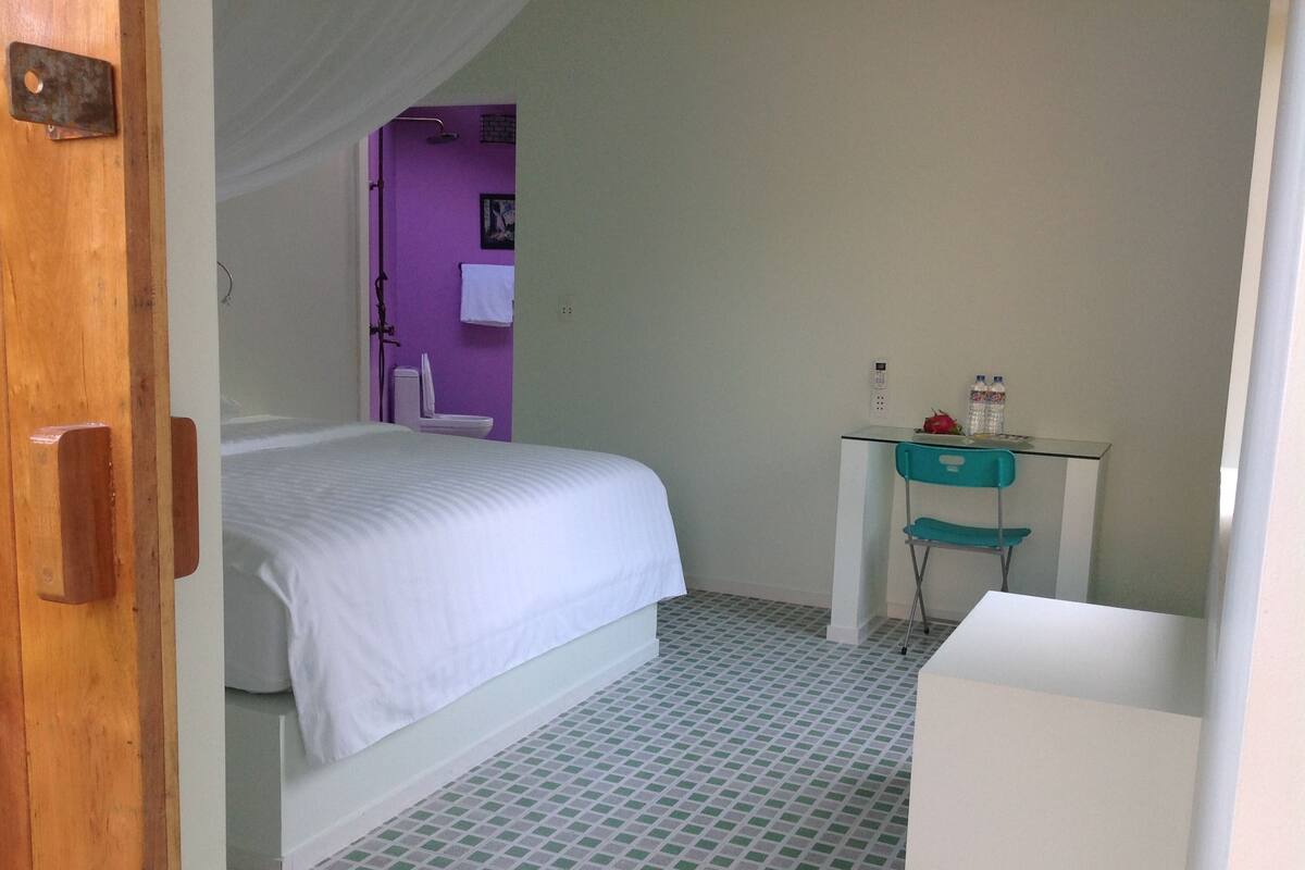 A bedroom with a bed, a small desk with a chair, a purple wall, and a tiled floor.