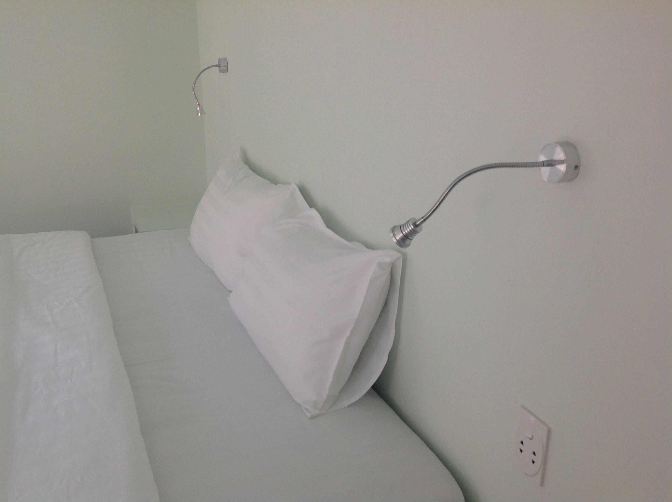 A bed with white bedding and a white pillow, a wall-mounted light fixture, and a power outlet.