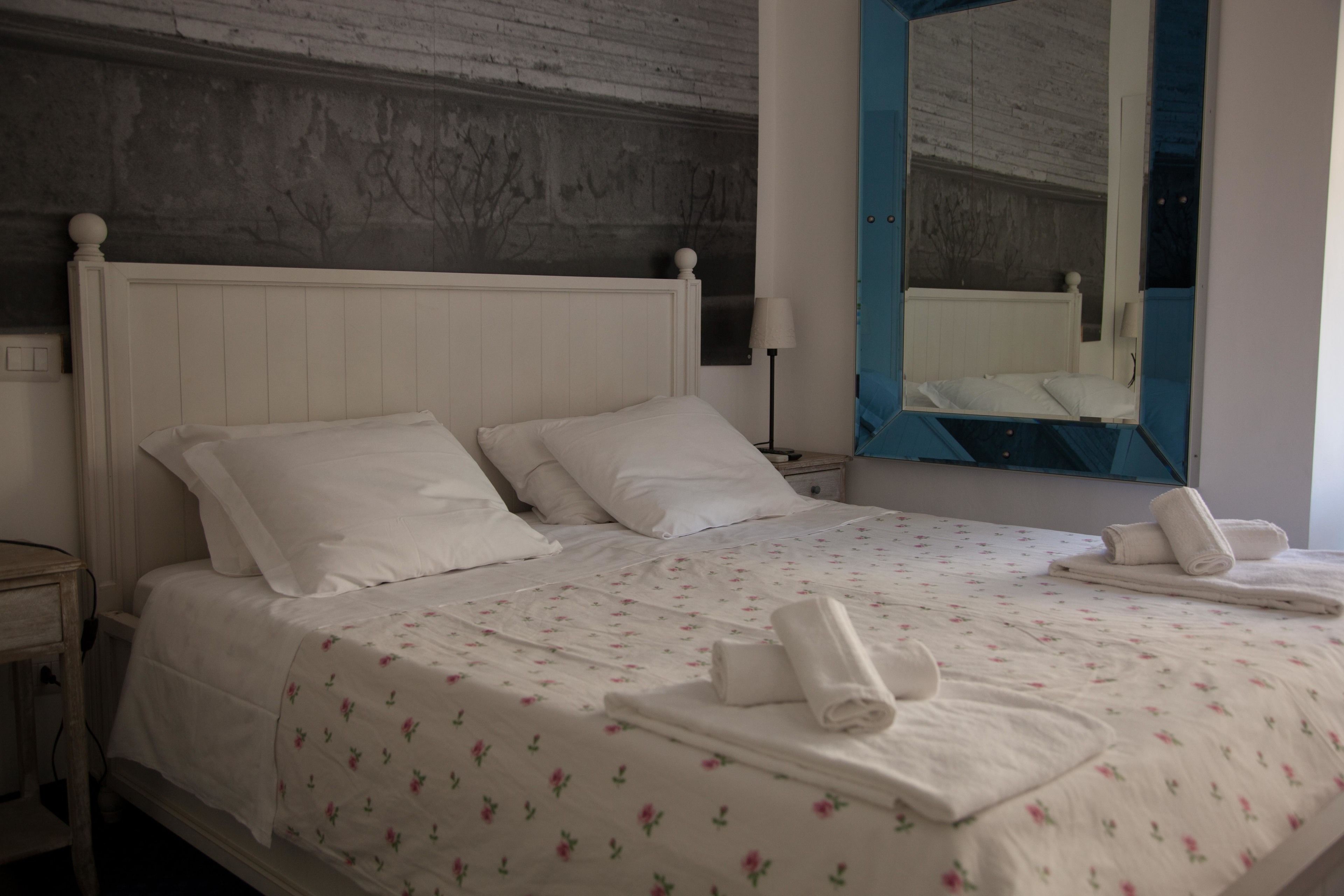 A neatly made bed with white linens and floral patterns, a wooden headboard, and a large mirror with a blue frame.