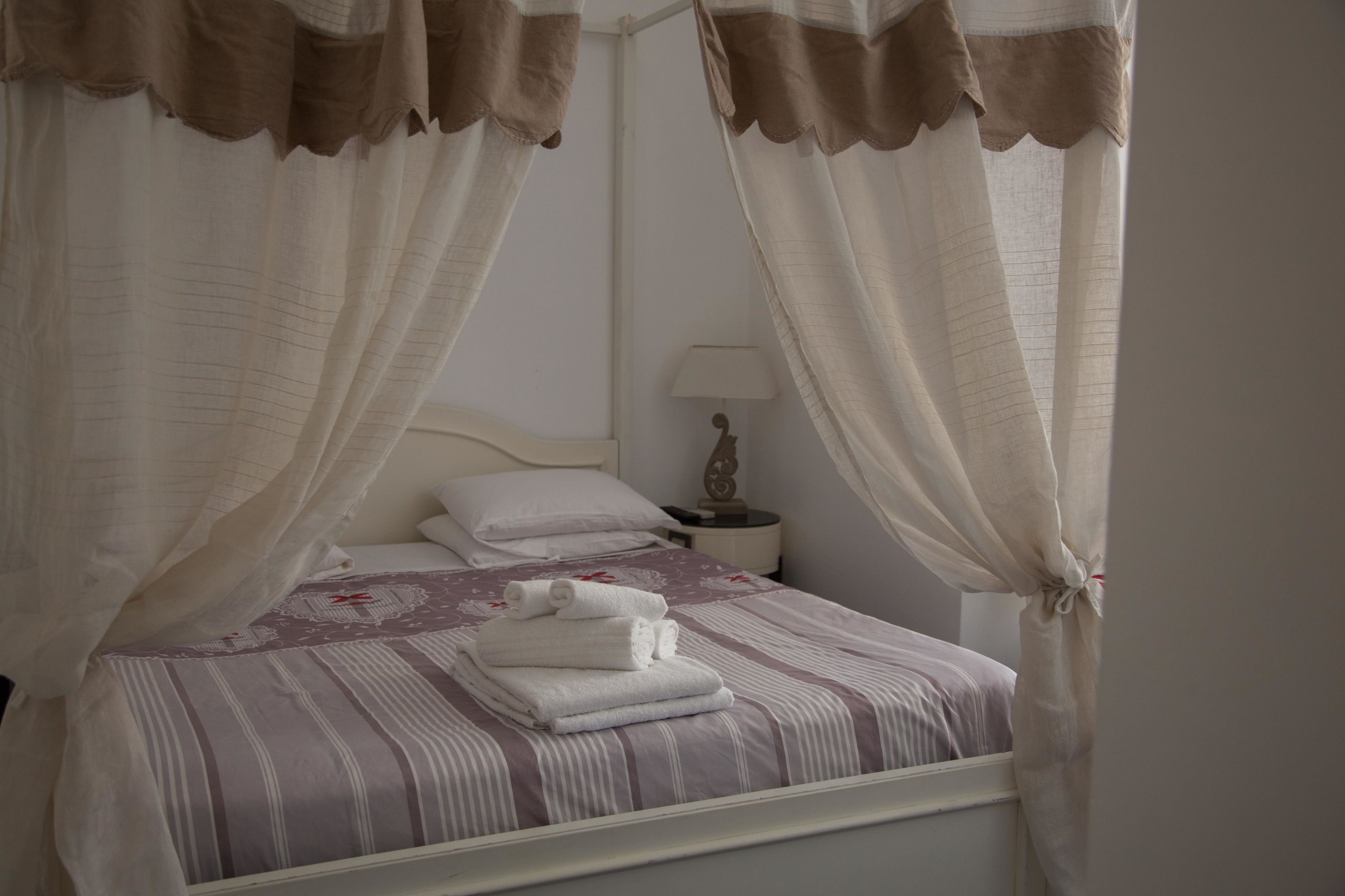 Una cama bien hecha con cubrecama a rayas y almohadas, una mesita de noche con lámpara, y cortinas traslúcidas con detalles marrones.
