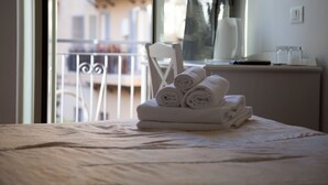 Una cama bien tendida con toallas enrolladas, una silla blanca y una ventana con vista a un balcón.