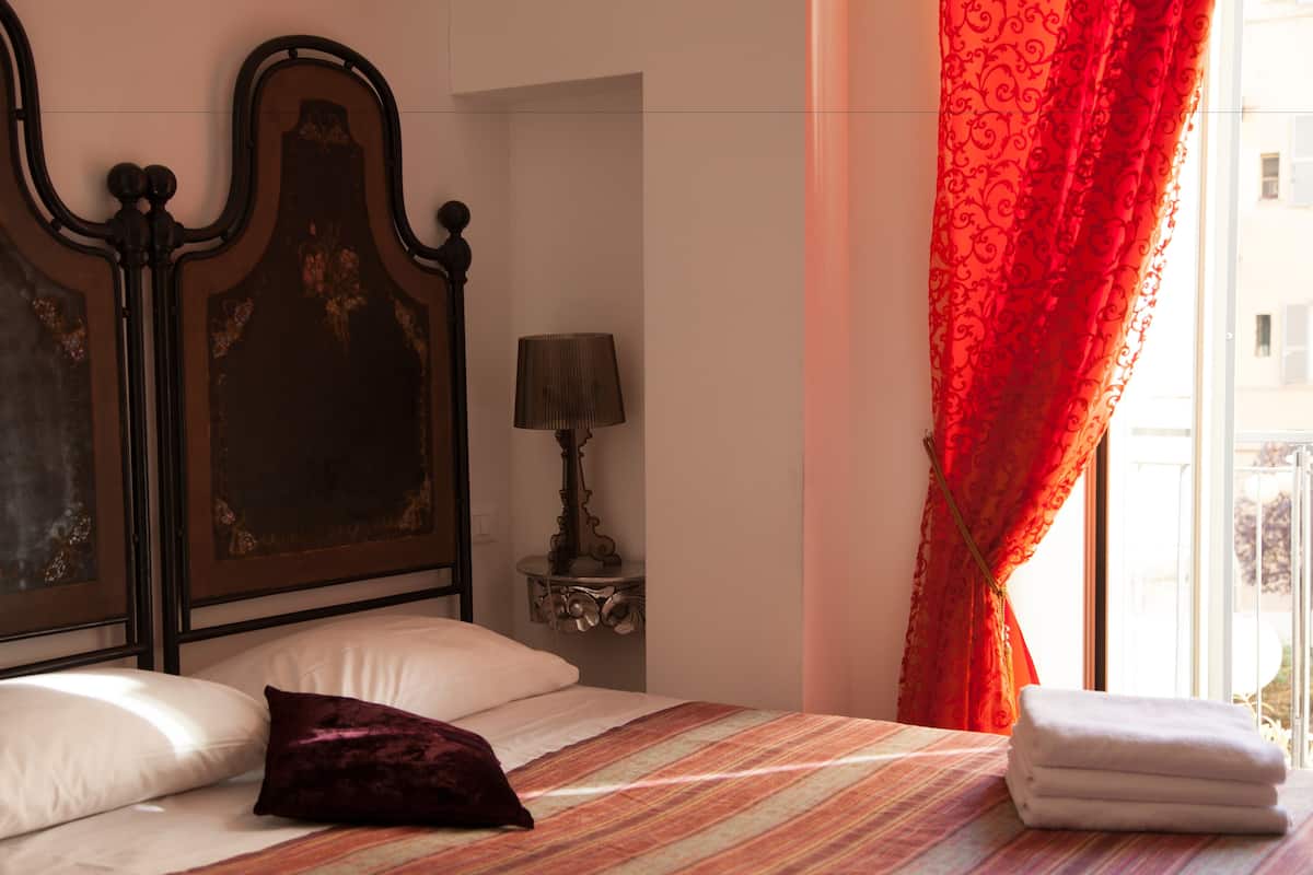 A neatly made bed with a striped bedspread, a dark headboard, a small bedside table with a lamp, and a window with red curtains.