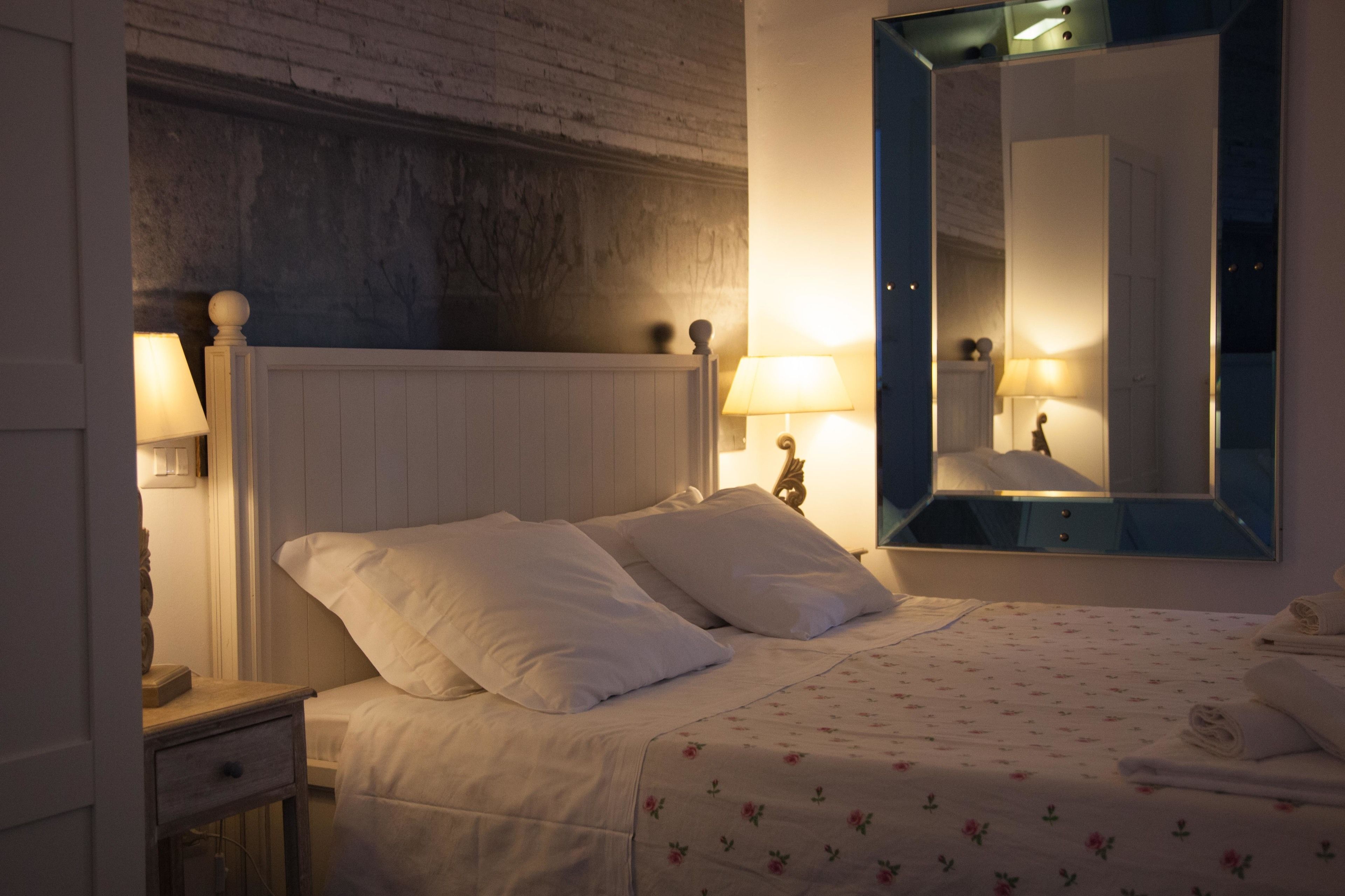 A neatly made bed with white linens and floral-patterned bedding, a wooden nightstand with a lamp, and a large mirror on the wall.