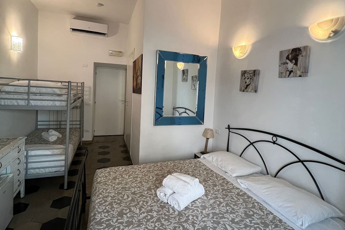 A hotel room with a bunk bed, a large bed with a patterned bedspread, a mirror, wall-mounted lights, and artwork.