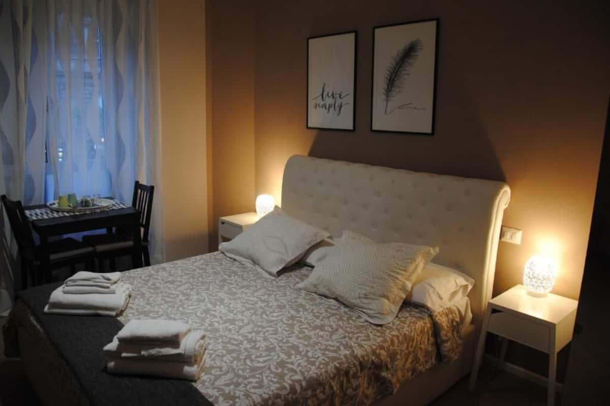 A neatly made bed with pillows and folded towels, a bedside table with a lamp, a chair, and a table with a lamp. Two framed pictures are on the wall.