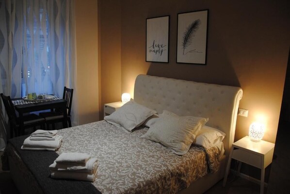 Un letto rifatto con cuscini e asciugamani piegati, un comodino con una lampada, una sedia e un tavolino anch'esso con lampada. Due quadri appesi al muro.