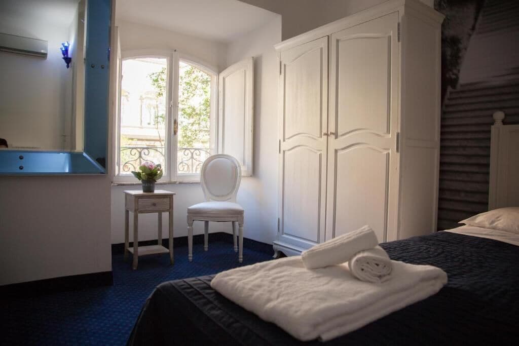 A hotel room with a bed, a chair, a desk, a wardrobe, and a window with a view of greenery.