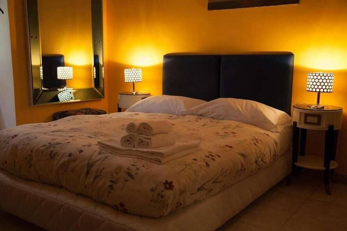 A bed with a floral bedspread, two bedside lamps, a mirror, and a folded towel on the bed.