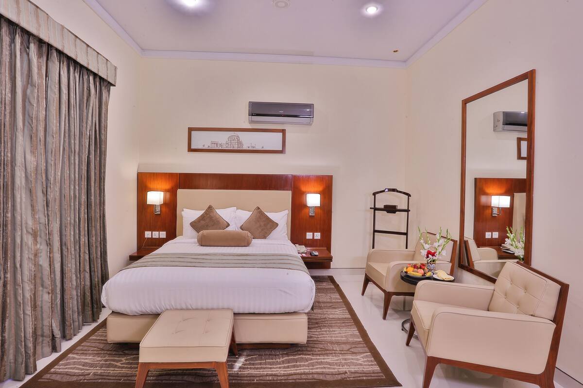 A hotel room with a large bed, bedside tables, a chair, a small table with fruit, and a mirror.