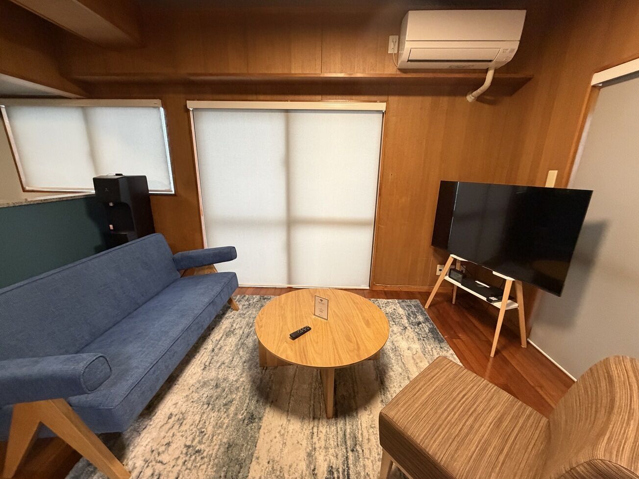 Une pièce avec un canapé bleu, une table ronde en bois, un téléviseur posé sur un meuble et une grande fenêtre avec des stores.