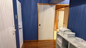 A room with a wooden floor, a white door, and a blue wall. There is a wooden wardrobe with a mirror and a shelf with folded towels. - Kokusai Towns Inn (Naha)