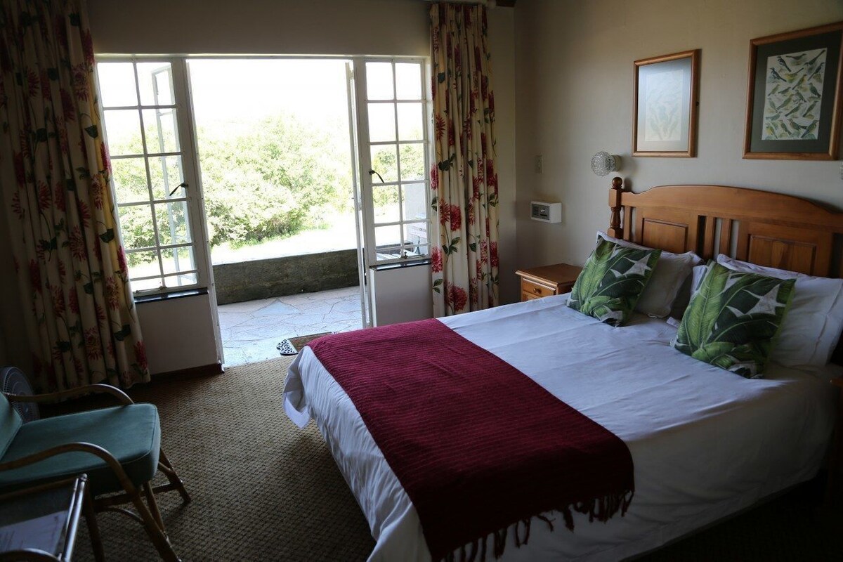 Una habitación con cama, una silla, un ventanal con cortinas y vistas al exterior.