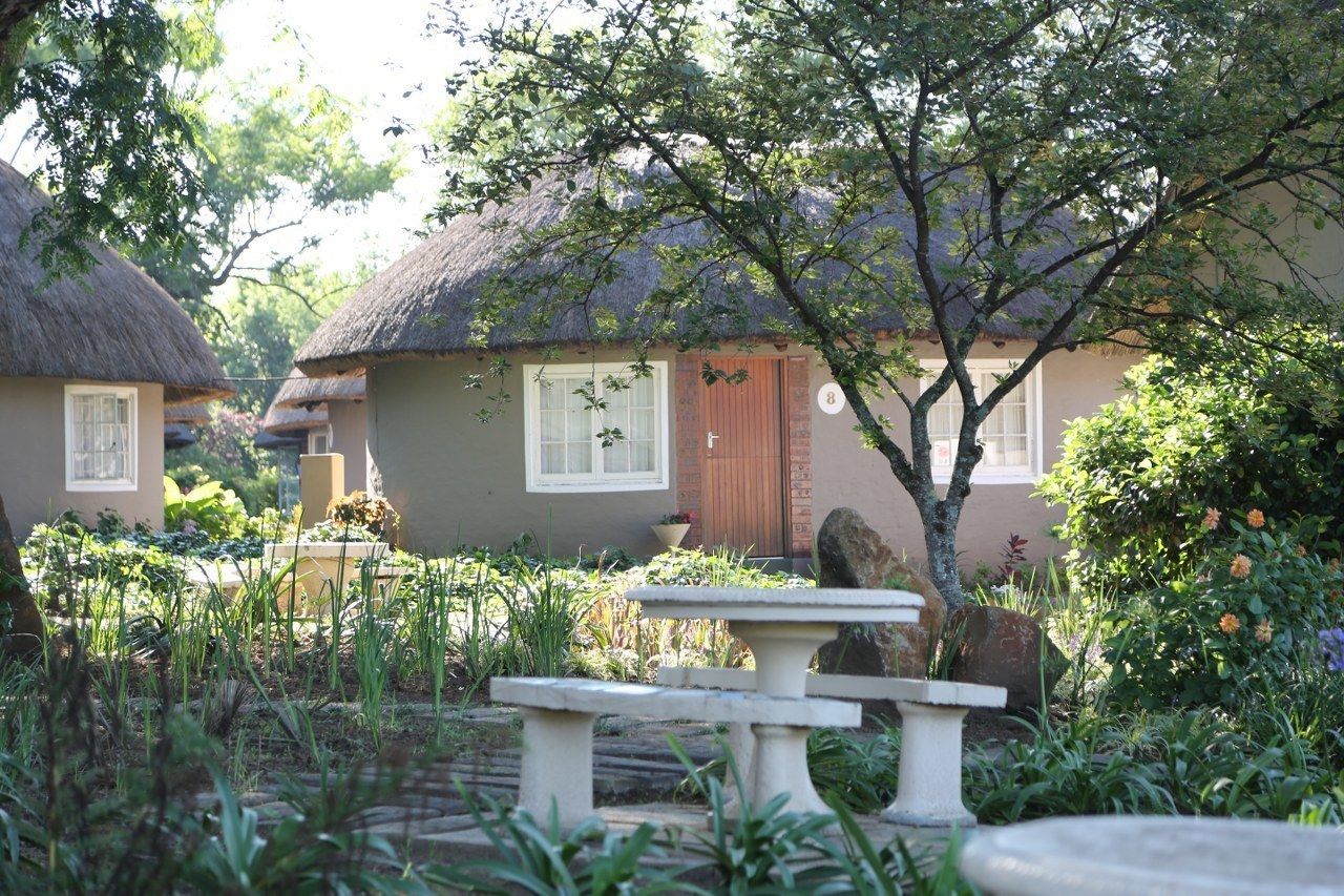 A traditional thatched-roof house with a garden in front, a stone bench, and a large tree.