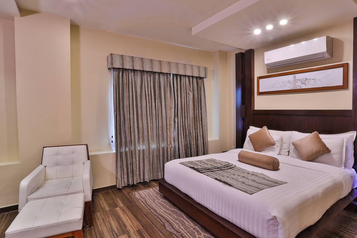 A hotel room with a bed, a chair, a window with curtains, an air conditioning unit, and a framed picture on the wall.