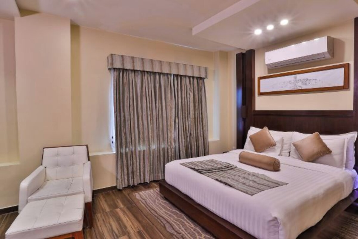 A hotel room with a bed, a chair, a window with curtains, an air conditioning unit, and a framed picture on the wall.