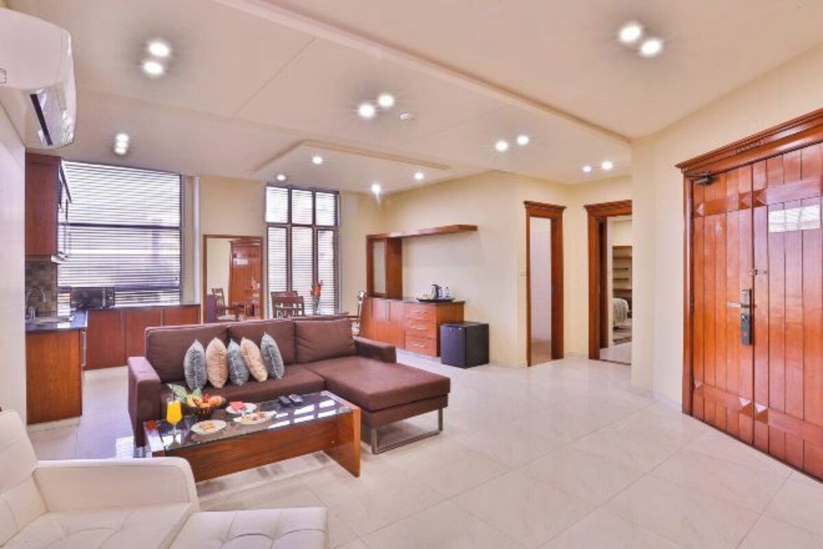 A spacious living area with a sofa, coffee table, and dining table set with food. There is a kitchen area with a refrigerator and cabinets. The room has wooden doors and a tiled floor.