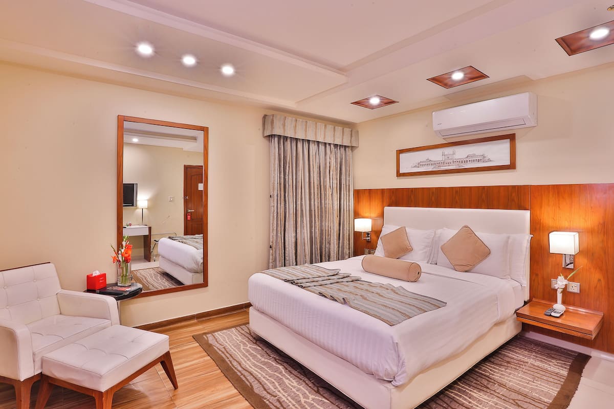 A hotel room with a bed, a chair, a mirror, a bedside table with a lamp, and an air conditioning unit.