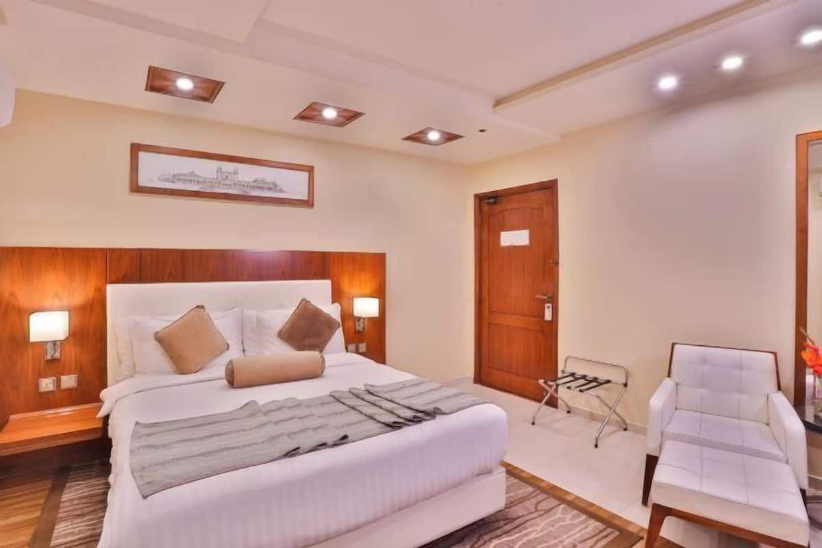A hotel room with a large bed, bedside lamps, a wooden headboard, a framed picture on the wall, a door, and a white armchair.