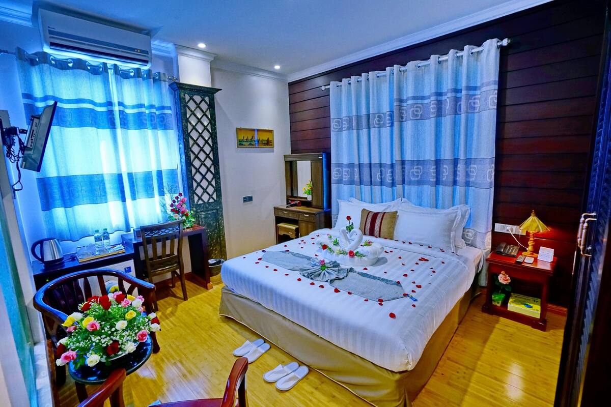 Habitación de hotel con una cama grande, un escritorio con silla, una mesita con lámpara y una mesita de noche con teléfono y una caja.