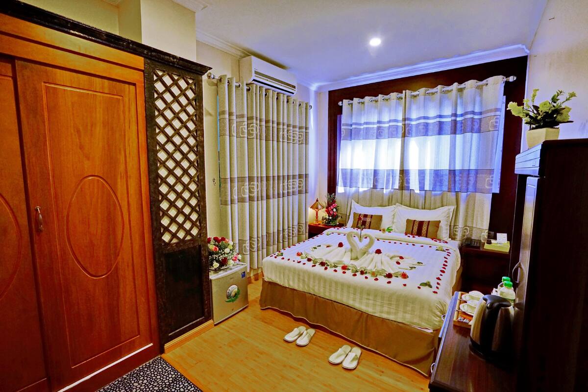 Habitación de hotel con armario de madera, cama con colcha blanca y adornos de rosa, mesita de noche con lámpara y flores, y ventana con cortinas.