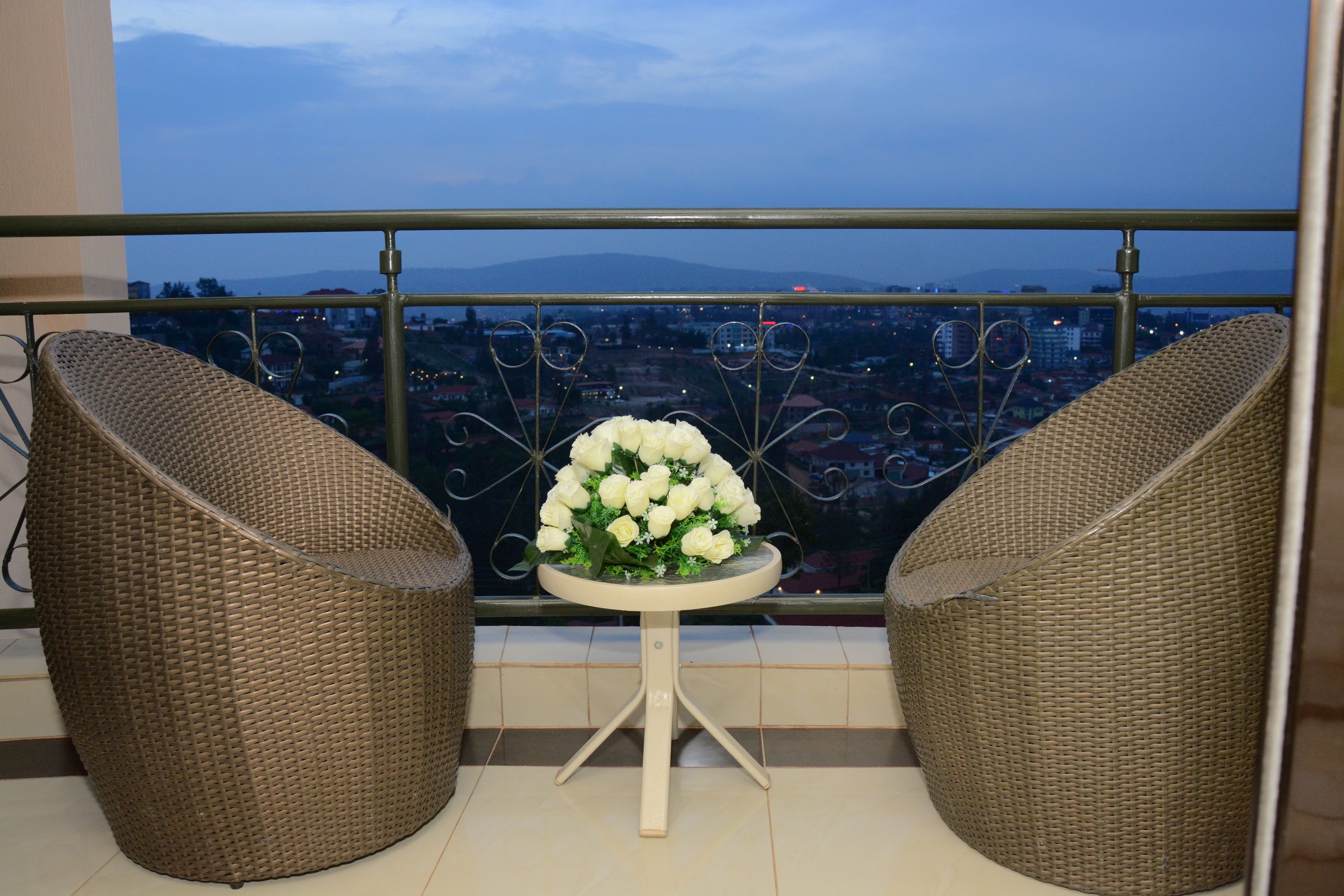 Ein Balkon mit Korbstühlen und einem Tisch mit einer weißen Blumenkomposition, mit Blick auf eine Stadtlandschaft.