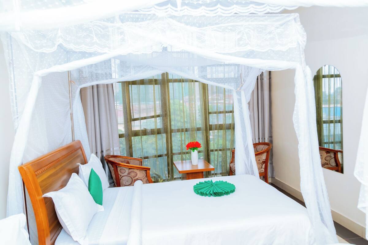 Una cama con dosel, una mesita con un florero y vistas a la vegetación a través de amplias ventanas.
