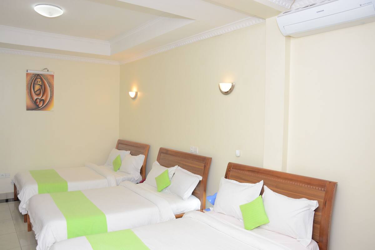 Habitación de hotel con tres camas, cada una con cabecero de madera, ropa de cama blanca y detalles en verde.