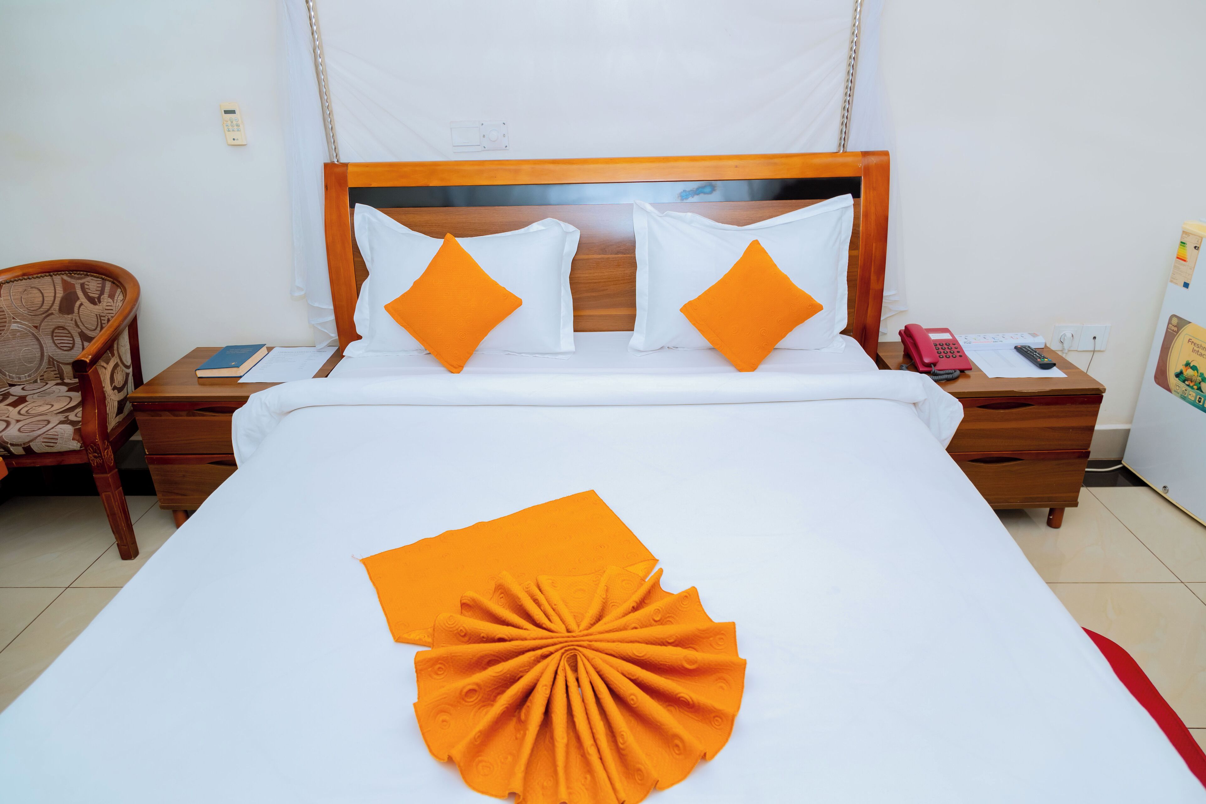Una cama bien hecha con una colcha blanca y naranja, dos mesitas de noche de madera con un teléfono rojo y un libro, y una silla con un cojín estampado.
