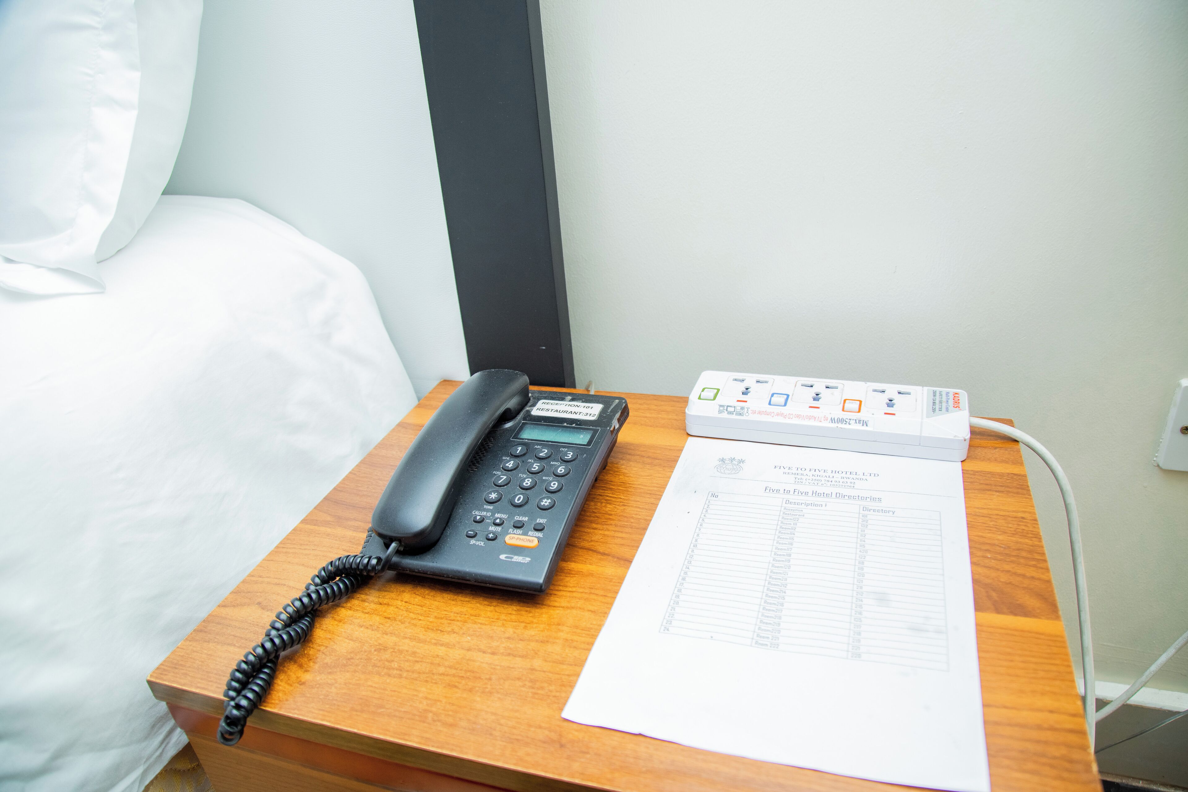 A wooden bedside table with a black telephone, a white hotel directory, and a white pillow on a bed.