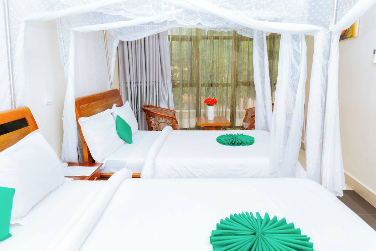 Una habitación de hotel con una cama, cabecera de madera, mesita de noche, una mesa con un florero y una ventana con cortinas translúcidas.